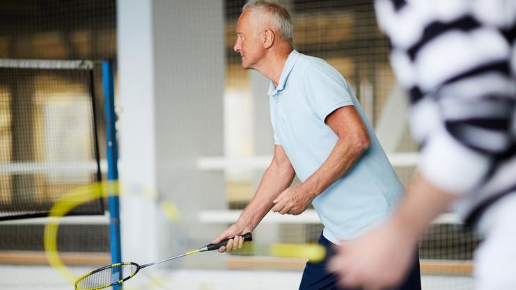 Älterer Mann mit Badminton-Schläger in der Hand, bereit zum Schlag auf einem Badmintonplatz. Hintergrund mit Netz sichtbar.