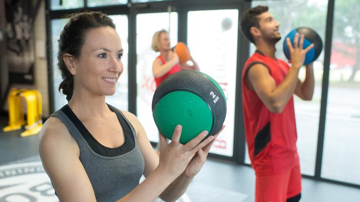 Drei Personen trainieren im Fitnessstudio mit Trainingsbällen; eine Frau hält einen grünen Medizinball.