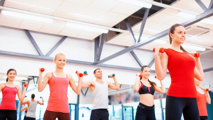 Gruppe von Menschen in Sportkleidung hebt Hanteln in einem Fitnessstudio mit Spiegeln und heller Beleuchtung.
