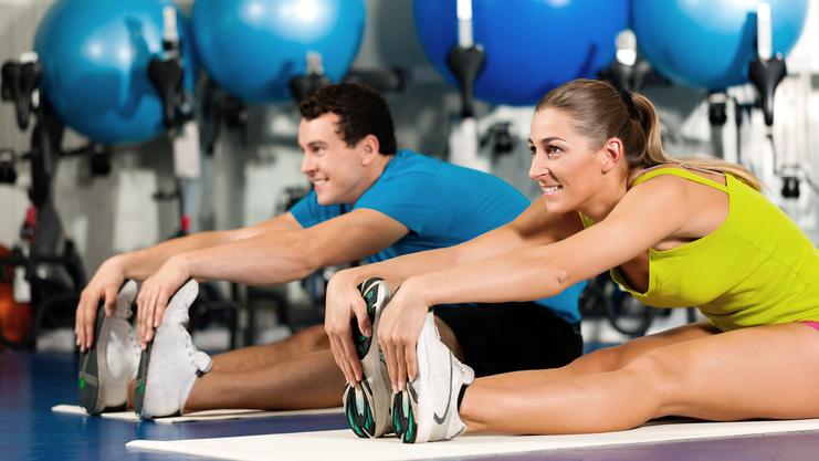 Zwei Personen dehnen auf Matten in einem Fitnessstudio, bunte Gymnastikbälle im Hintergrund.