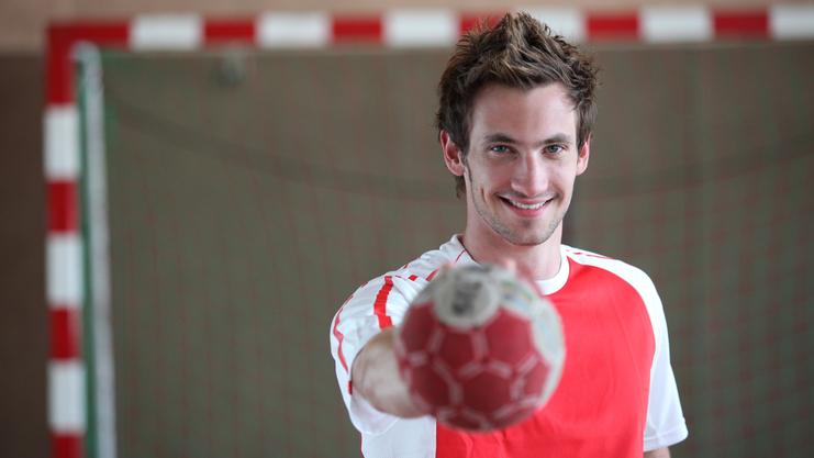 Junger Mann im roten Trikot hält einen Handball vor sich, lächelt in einer Sporthalle mit Tor im Hintergrund.