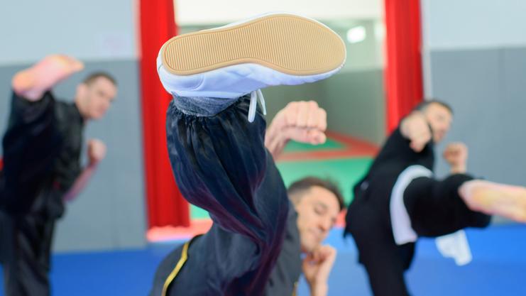 Drei Kampfsportler in Uniform üben Tritte auf einer blauen Matte in einem Trainingsraum mit roten Vorhängen.