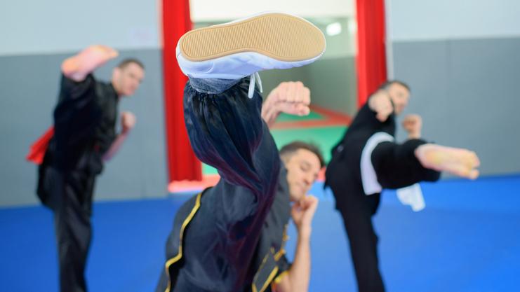 Drei Kampfsportler in Uniform üben Tritte auf einer blauen Matte in einem Trainingsraum mit roten Vorhängen.