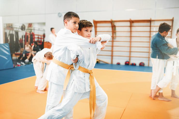 Zwei Jungen in weißen Judoanzügen üben eine Technik auf einer Judo-Matte, während andere im Hintergrund trainieren.