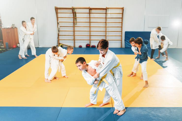 Gruppe von Kindern in Judokleidung trainiert auf einer gelben und blauen Judomatte in einem Sportstudio.