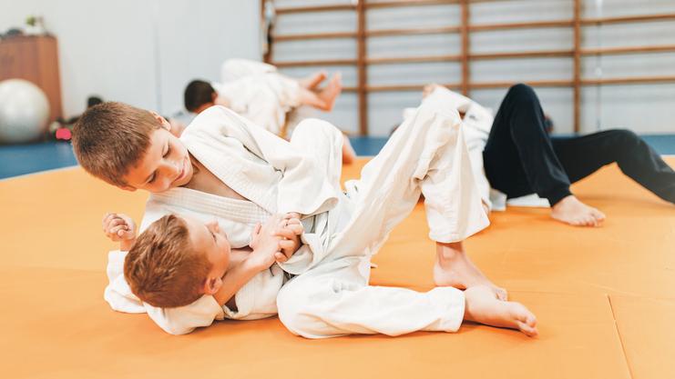 Zwei Kinder im Judo-Anzug üben auf einer orangefarbenen Matte, während andere im Hintergrund trainieren.