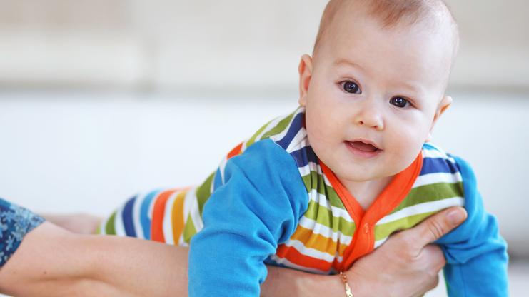 Baby in bunten Strampelanzug liegt auf einem grünen Gymnastikball, von einer Hand unterstützt.