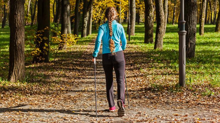 Frau mit langem Zopf geht mit Wanderstöcken einen herbstlichen Weg zwischen Bäumen, umgeben von buntem Laub.