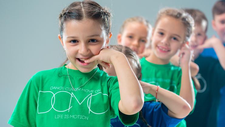 Lächelnde Kinder in bunten T-Shirts machen eine Tanzbewegung vor einem hellen Hintergrund.