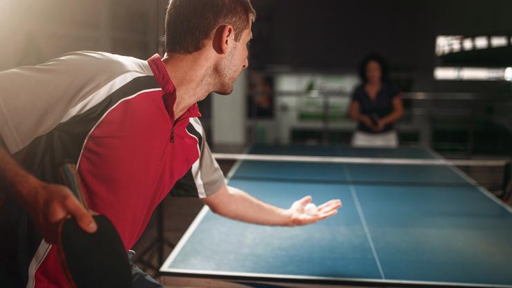 Mann in rot-schwarzem Shirt bereitet sich auf einen Aufschlag am Tischtennistisch vor, während eine Frau zusieht.